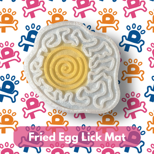 A fried egg-shaped lick mat with raised patterns, designed for dogs to lick treats off. The mat is presented on a background with colorful pet-themed graphics.