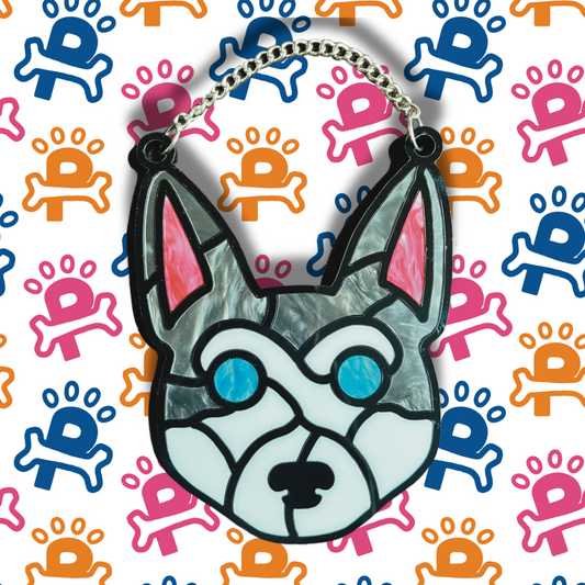 A pho stained glass decoration depicting a husky's face, crafted with intricate pieces of colored acrylic. The design features the husky's signature markings in shades of white, gray, and black, with striking blue for the eyes. The decoration is framed in a thin black outline and hangs by a delicate chain.